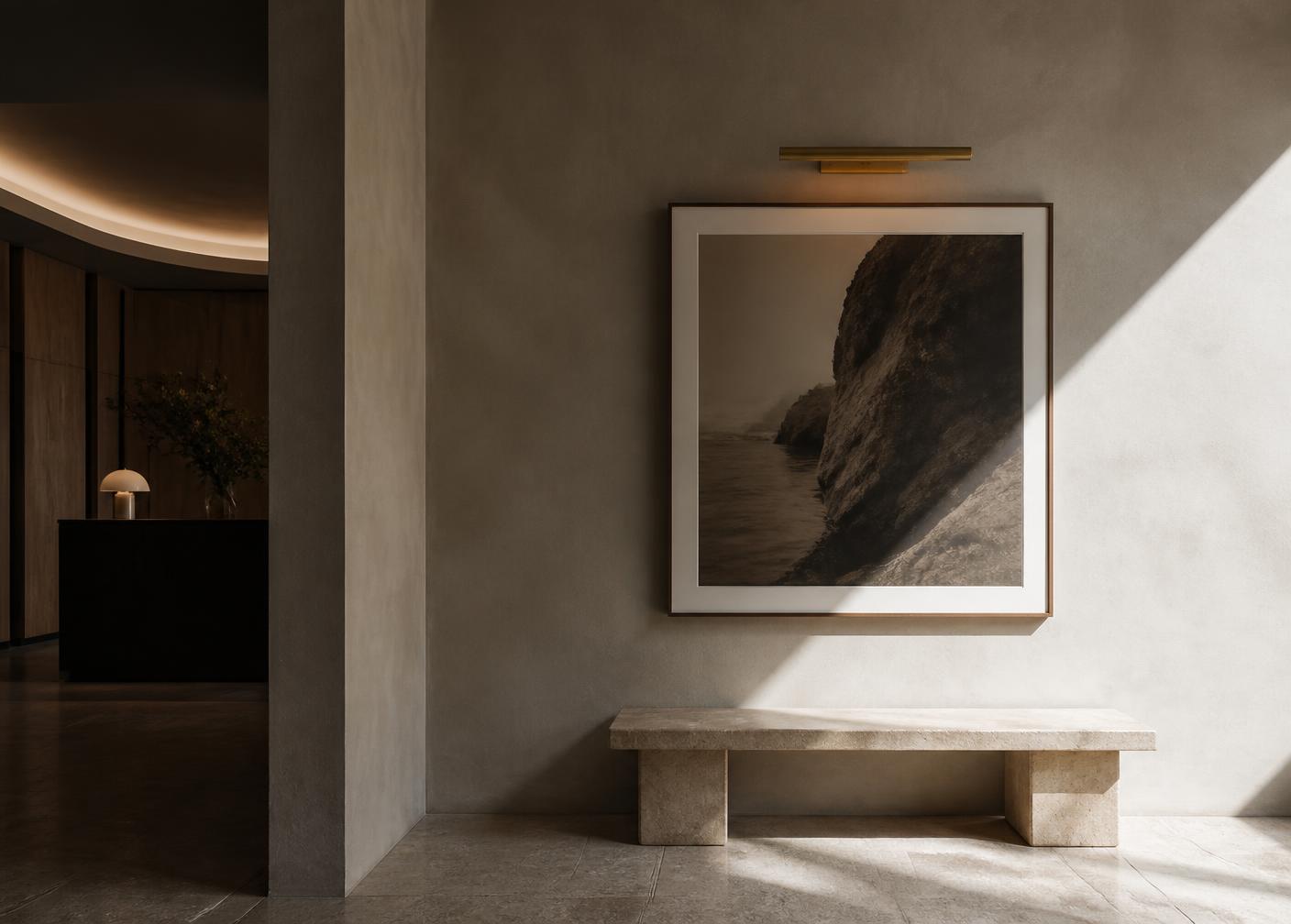 A framed photographic artwork lit by a slim brass wall light above a stone bench in a boutique hotel lobby